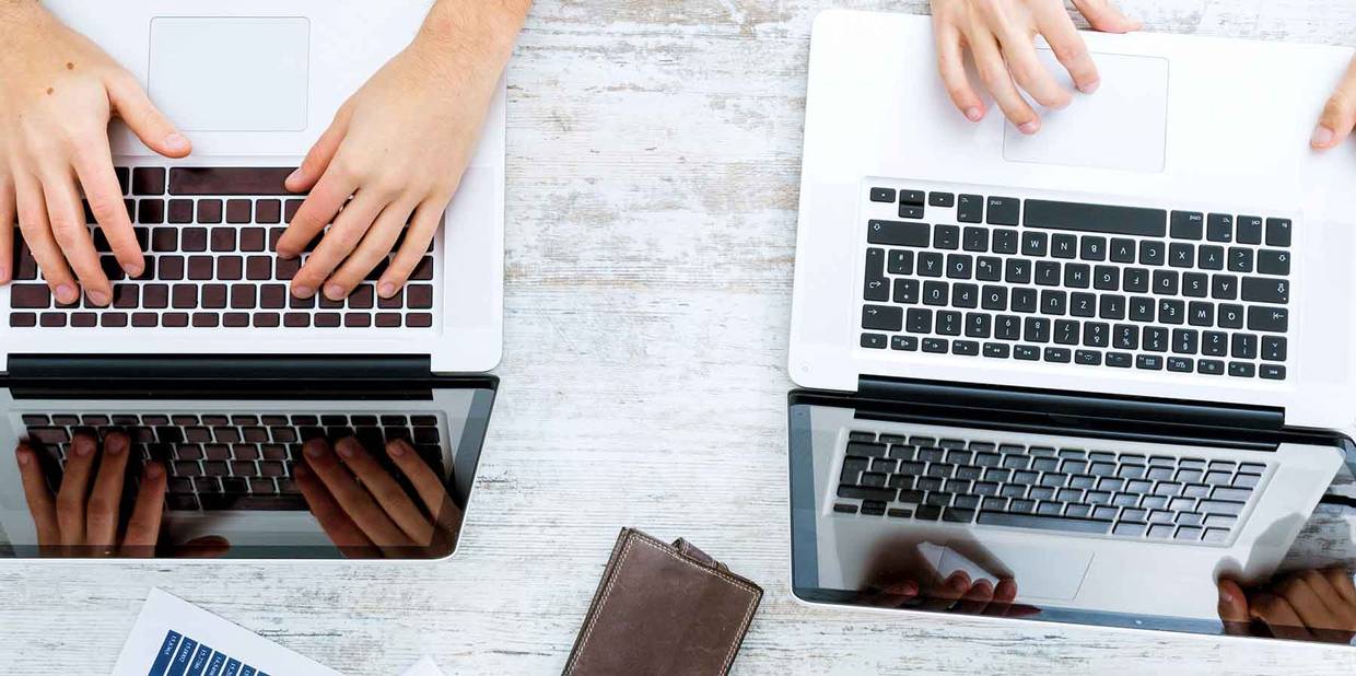 Dois computadores estão em cima de uma bancada de madeira branca e duas pessoas trabalham neles