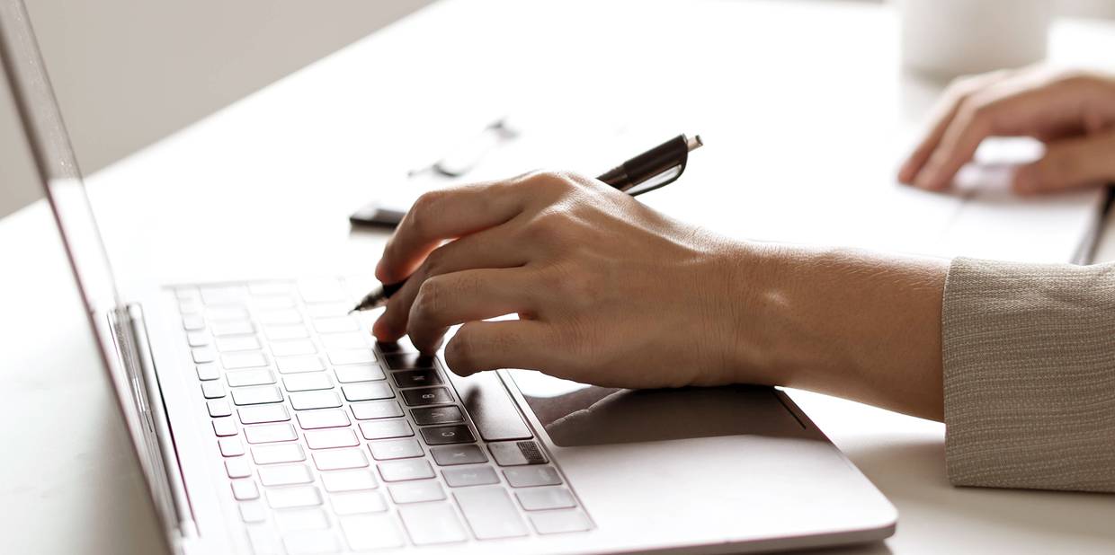 Um notebook aparece em cima de uma mesa e mãos de uma pessoa estão posicionadas no aparelho