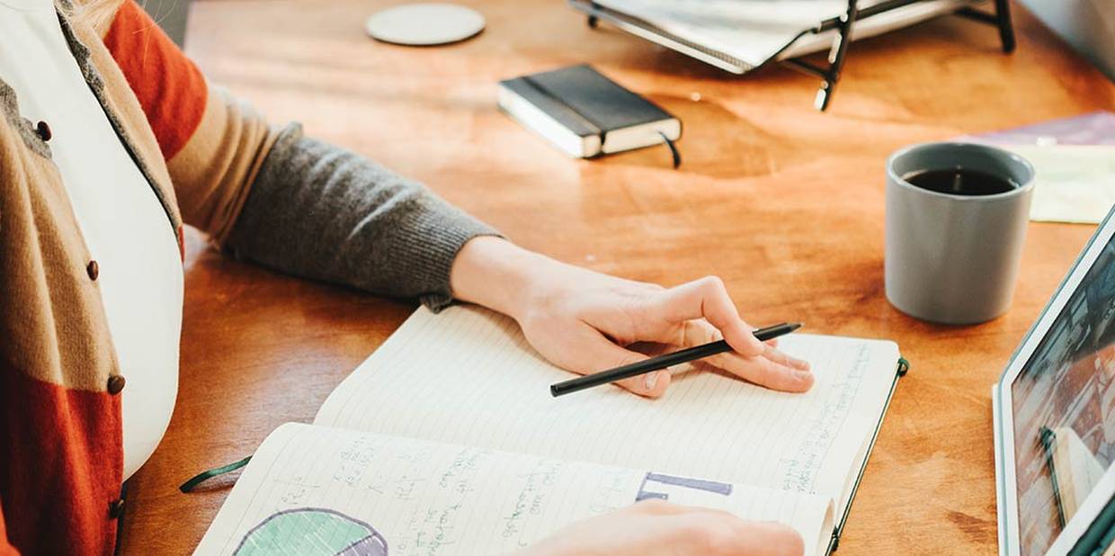 Foto colorida mostra mãos sobre um caderno que está em cima de uma mesa. Na mesa há porta-retrato, xícara e um bloco de notas.