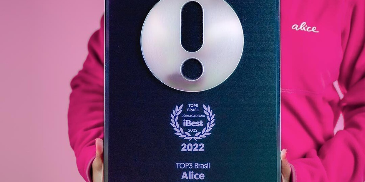 Torso de uma pessoa vestindo um moletom rosa da Alice enquanto segura nas mãos o prêmio iBest. O prêmio iBest 2022 sendo segurado é da categoria TOP3 Brasil Healthtech - Alice .