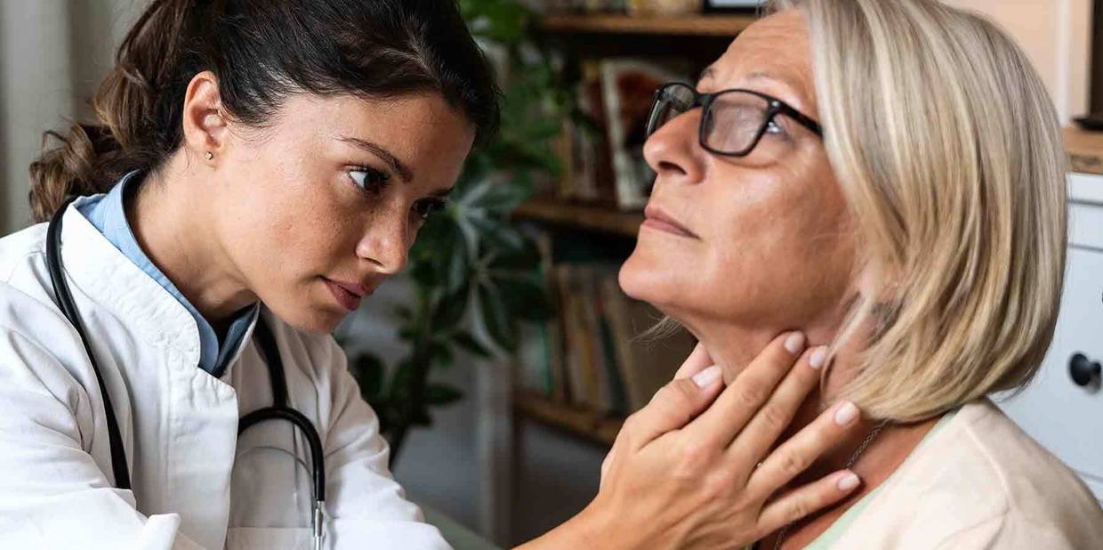 Médica branca apalpa o pescoço de paciente, uma mulher branca a sua frente