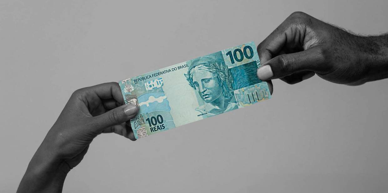 Uma nota de 100 reais está em destaque enquanto duas mãos seguram o dinheiro, uma em cada ponta