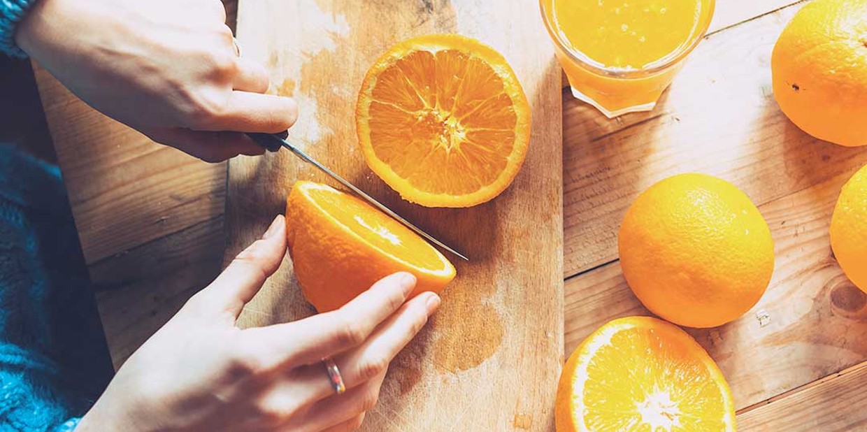 Close das mãos de uma mulher cortando laranjas, um dos alimentos que podem piorar a afta