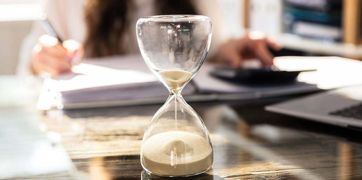 Ampulheta de areia virada e o tempo está se encerrando. No fundo vemos uma mulher escrevendo em um caderno com uma caneta e mexendo em uma calculadora.