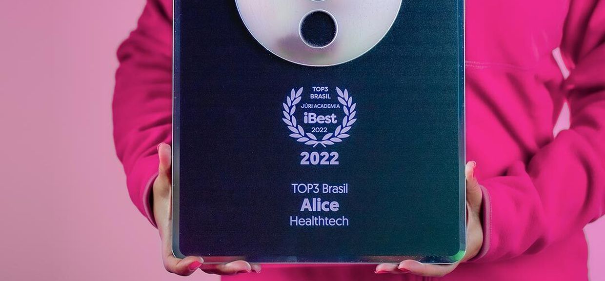 Torso de uma pessoa vestindo um moletom rosa da Alice enquanto segura nas mãos o prêmio iBest. O prêmio iBest 2022 sendo segurado é da categoria TOP3 Brasil Healthtech - Alice .