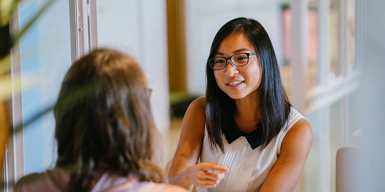 Duas mulheres conversando em uma entrevista de emprego. Uma das mulheres tem descendência oriental, usa óculos e uma regata branca e tem cabelo preto. A outra está de costas e usa uma regata rosa.