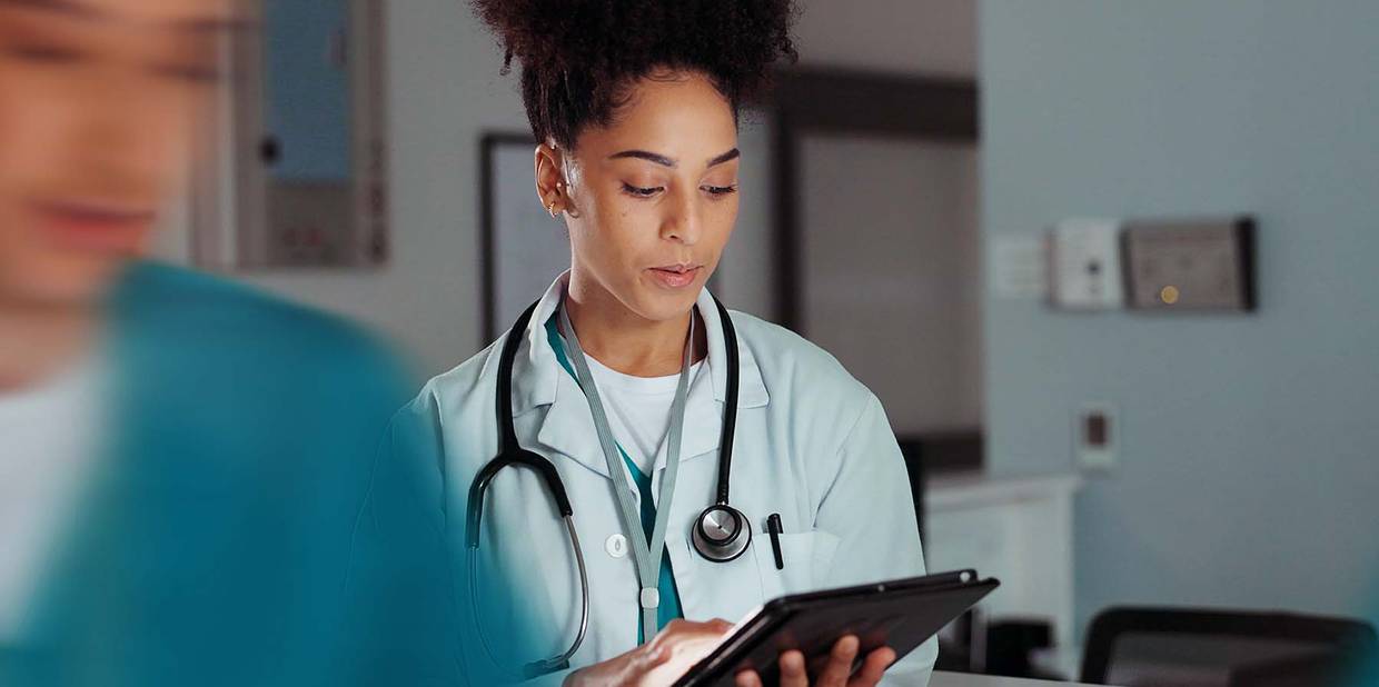 Uma mulher médica está com jaleco e materiais de trabalho em um ambiente hospitalar com um tablet na mão