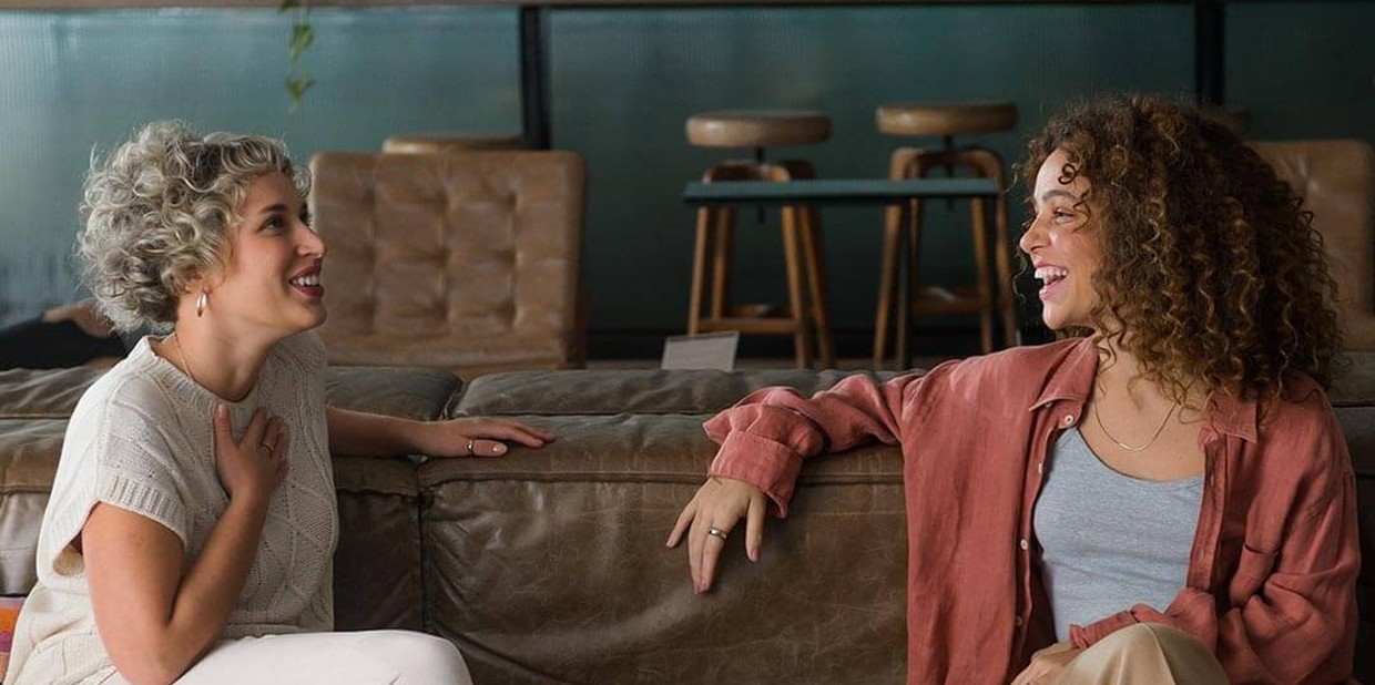 Duas mulheres sentadas riem sentadas em um sofá de couro marrom.