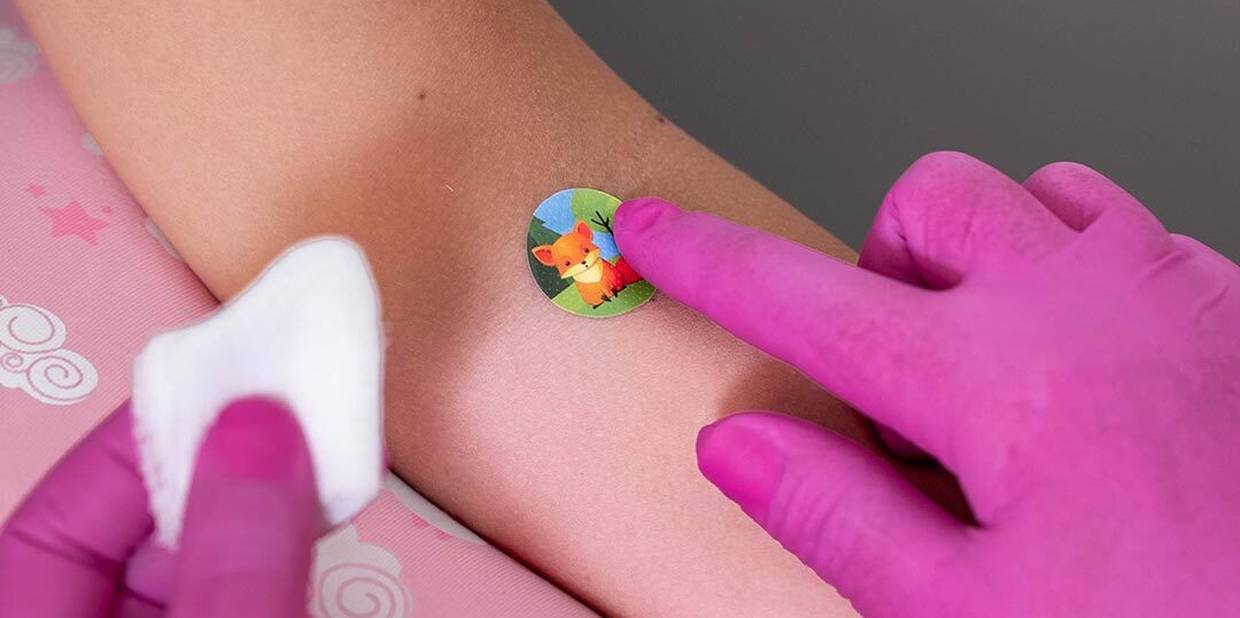 Close do braço de uma pessoa com uma enfermeira colocando um curativo sobre o local da coleta de sangue. O curativo tem uma ilustração de uma raposinha.