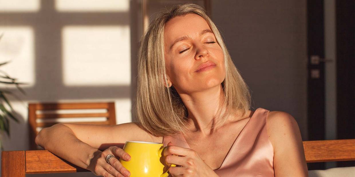 Uma mulher está sentada com uma xícara na mão e relaxa enquanto recebe o sol no rosto