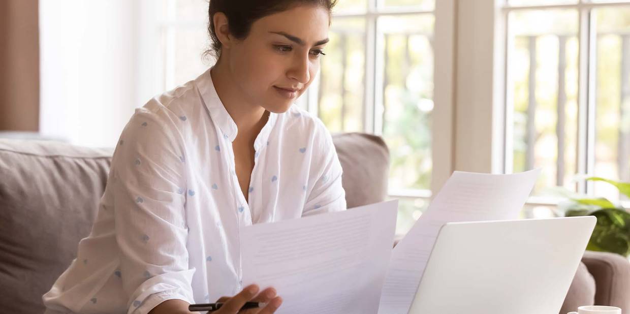 Uma mulher sentada com um computador a sua frente analisa folhas de papel enquanto trabalha