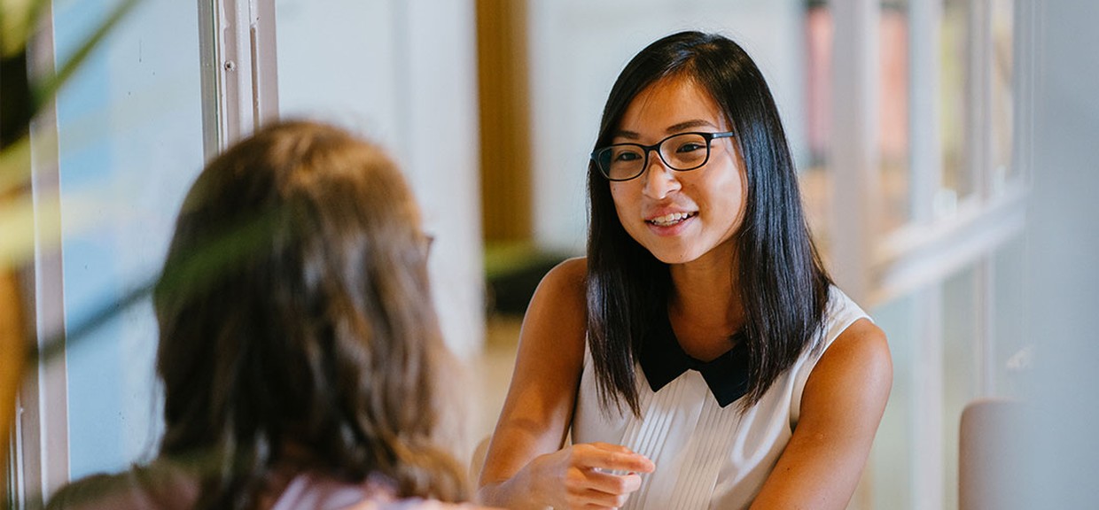 Duas mulheres conversando em uma entrevista de emprego. Uma das mulheres tem descendência oriental, usa óculos e uma regata branca e tem cabelo preto. A outra está de costas e usa uma regata rosa.