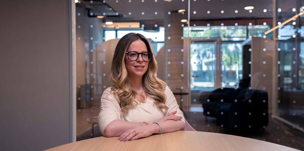Marcia Denes está sentada com os braços em cima de uma mesa de madeira. Ela tem cabelos loiros, usa óculos e está com uma roupa branca