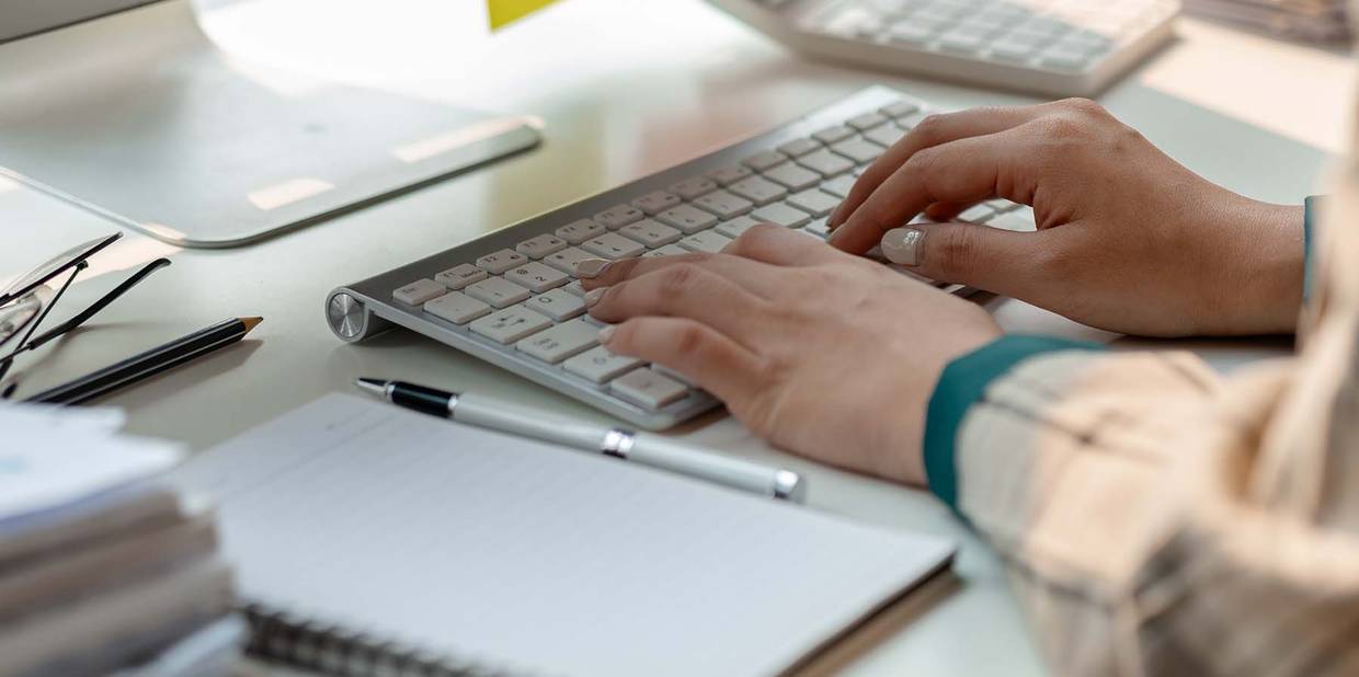 Uma mesa branda tem um computador e outros objetos de escritório. Uma pessoa está com as duas mãos no teclado do computador